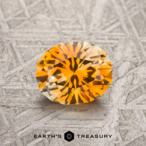 A 1.29-carat orange-yellow particolored heated Montana sapphire is displayed on a textured metallic surface with "EARTH'S TREASURY" at the bottom.
