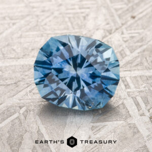 A 1.74-carat sky blue Montana Sapphire (heated), cut in a round brilliant style, is displayed on a textured surface with the text "Earth's Treasury" below it.