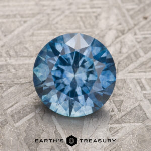 A 1.69-carat medium blue Montana Sapphire (heated), round and faceted, is shown upright on a patterned metallic surface, with the "EARTH'S TREASURY" logo and text below it.