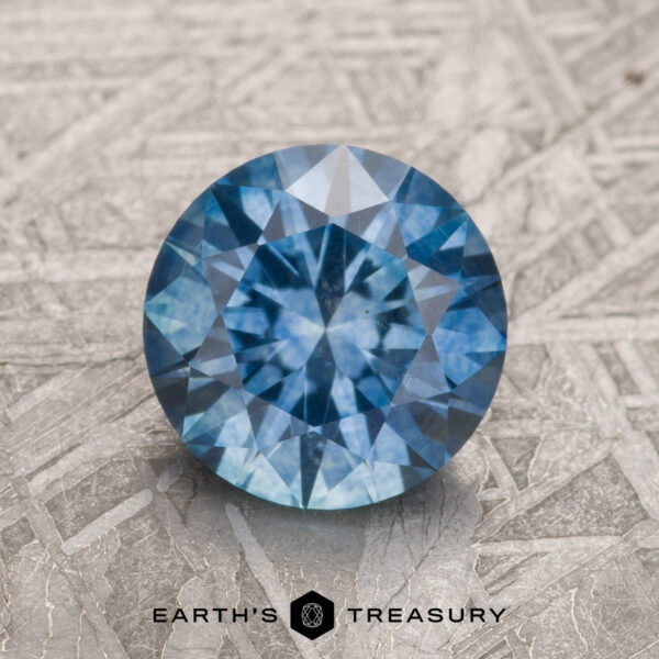1.69-Carat Medium Blue Montana Sapphire (Heated) A 1.69-carat medium blue Montana Sapphire (heated), round and faceted, is shown upright on a patterned metallic surface, with the "EARTH'S TREASURY" logo and text below it.