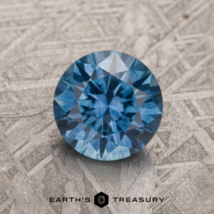 A 1.50-carat Rich Teal Montana Sapphire (Heated), round and faceted, is displayed on a textured gray surface with "EARTH'S TREASURY" and a logo at the bottom.