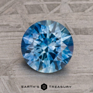 A 1.30-carat Medium Blue Montana Sapphire (Heated), round brilliant-cut, is displayed on a textured gray surface with the Earth’s Treasury logo beneath it.