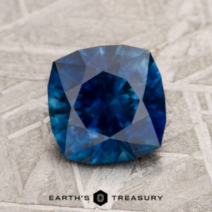 A 1.92-carat Deep Blue Montana Sapphire (heated) with cushion cut and geometric facets rests on a textured metallic surface, with the Earth’s Treasury logo at the bottom of the image.