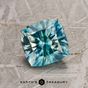 A 1.95-carat blue-green-yellow particolored Montana sapphire (heated) with multifaceted reflections is displayed on a textured gray surface. The Earth’s Treasury logo is visible at the bottom.