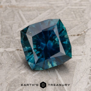 A 1.81-carat deep blue-green heated Montana Sapphire is displayed on a textured gray surface with "EARTH'S TREASURY" written below.