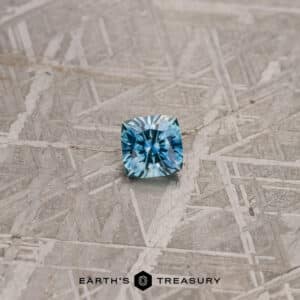 A 0.86-carat teal blue Montana sapphire with intricate faceting lies on a textured surface resembling etched metal. The words "EARTH'S TREASURY" are displayed at the bottom of the image.