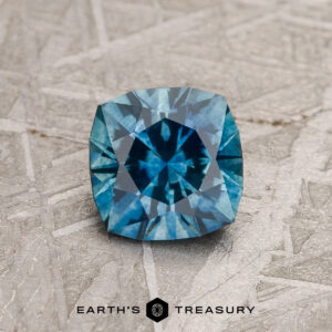 A 1.06-carat Teal Blue Montana Sapphire (heated), cushion-cut with intricate facets, sits on a textured gray surface. "EARTH'S TREASURY" is displayed at the bottom of the image.