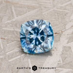 A 1.03-carat Sky Blue Montana Sapphire (heated) with a cushion cut and starburst pattern rests on a textured gray surface, with the Earth's Treasury logo visible at the bottom.