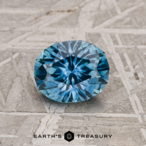 A 1.27-carat heated teal blue Montana sapphire is showcased on a textured gray surface with the “Earth’s Treasury” logo and text at the bottom.