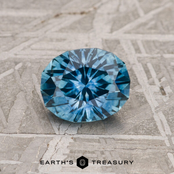 1.27-Carat Teal Blue Montana Sapphire (Heated) A 1.27-carat heated teal blue Montana sapphire is showcased on a textured gray surface with the “Earth’s Treasury” logo and text at the bottom.