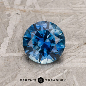 A 1.14-carat Medium Blue Montana Sapphire (Heated), round brilliant-cut, is shown on a textured gray surface with the "EARTH'S TREASURY" logo and text at the bottom.