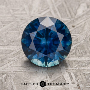 A 2.06-carat deep blue heated Montana Sapphire, round brilliant-cut, is shown on a textured metallic surface with the "Earth's Treasury" logo and text at the bottom.