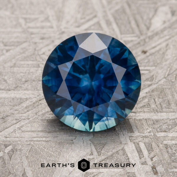 2.06-Carat Deep Blue Montana Sapphire (Heated) A 2.06-carat deep blue heated Montana Sapphire, round brilliant-cut, is shown on a textured metallic surface with the "Earth's Treasury" logo and text at the bottom.