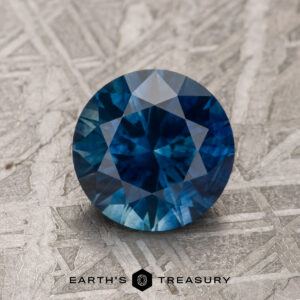 A 1.76-carat deep blue Montana Sapphire (heated), round and faceted, rests on a textured grey surface with the "EARTH'S TREASURY" logo and text below.