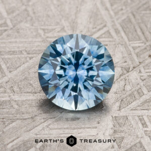 1.37-Carat Sky Blue Montana Sapphire (Heated) in a round brilliant cut, displayed on a metallic textured surface with the "Earth's Treasury" logo and text below the gemstone.
