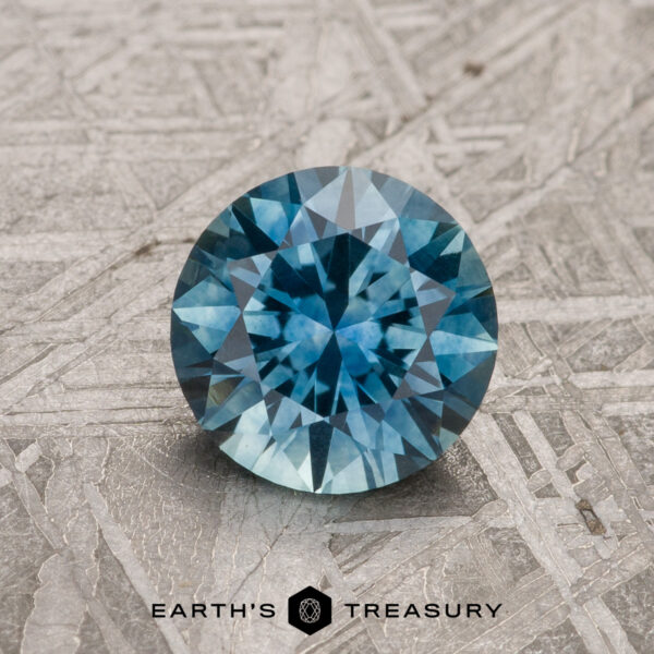 1.30-Carat Teal Blue Montana Sapphire (Heated) A 1.30-carat Teal Blue Montana Sapphire (heated), round and faceted, rests on a textured gray surface with "EARTH'S TREASURY" displayed at the bottom of the image.