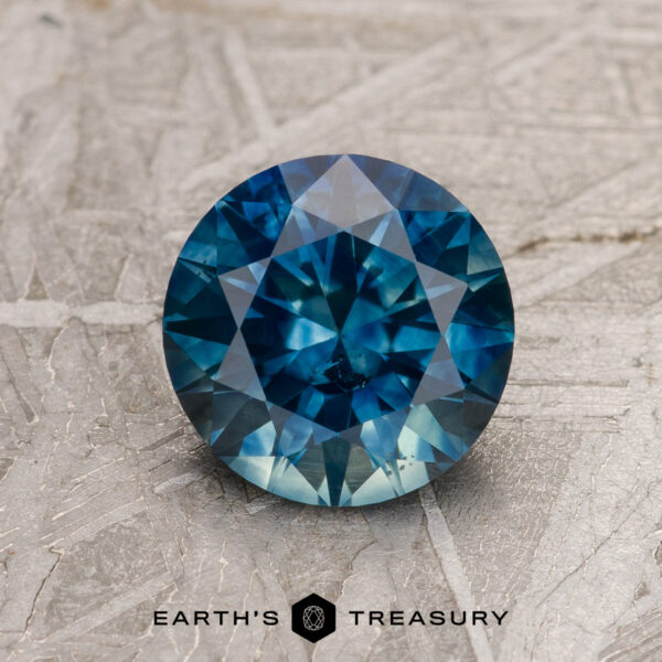 1.55-Carat Peacock Blue Montana Sapphire (Heated) A 1.55-carat peacock blue Montana Sapphire (heated), round brilliant-cut, is displayed on a textured gray surface with the text "EARTH'S TREASURY" beneath it.