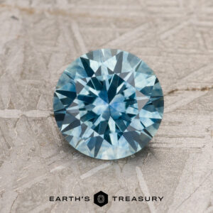 A 1.39-carat aqua blue Montana Sapphire (heated), round brilliant-cut, is displayed on a textured gray surface with "EARTH'S TREASURY" at the bottom.