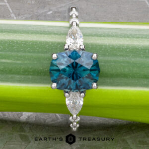 The "Carina" ring in 14k white gold, featuring a 1.53-carat Montana Sapphire flanked by marquise-cut gems, is displayed on a green plant stem. The "Earth's Treasury" logo appears at the bottom, enhancing its elegance.