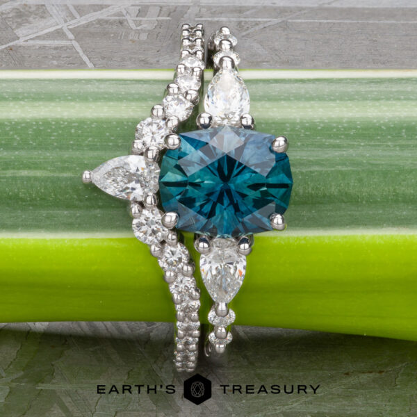 The "Carina" ring in 14k white gold with 1.53-Carat Montana Sapphire A 1.53-carat Montana Sapphire "Carina" ring in 14k white gold with diamond accents and a matching diamond band, both displayed on a green stem. "Earth's Treasury" logo appears at the bottom.