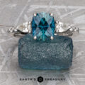 The "Carina" ring in 14k white gold, featuring a 1.53-carat Montana sapphire and two small clear stones, is showcased on a textured blue rock with the "Earth's Treasury" logo beneath.
