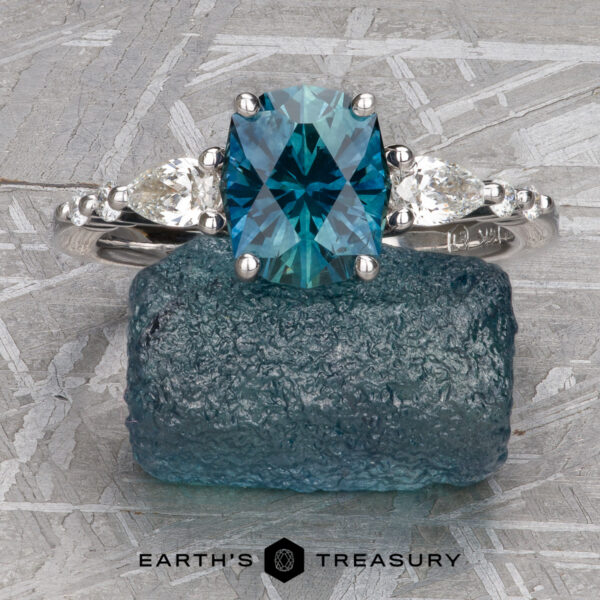 The "Carina" ring in 14k white gold with 1.53-Carat Montana Sapphire The "Carina" ring in 14k white gold, featuring a 1.53-carat Montana sapphire and two small clear stones, is showcased on a textured blue rock with the "Earth's Treasury" logo beneath.