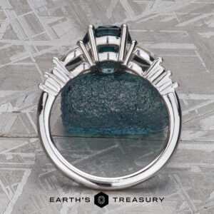 A "Carina" ring in 14k white gold with a 1.53-carat Montana sapphire is shown upright from the side on a textured surface, featuring the "Earth’s Treasury" logo at the bottom.
