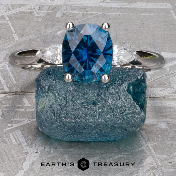The Minimal "Avalon" ring in 18k white gold with 1.69-Carat Montana sapphire The Minimal "Avalon" ring in 18k white gold with a 1.69-carat Montana sapphire and two clear side stones is displayed on rough blue crystal, with the "Earth's Treasury" logo below.