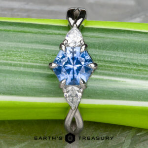 A pendant inspired by the "Elowen" ring in platinum, which features a 1.09-carat Montana sapphire at its center, flanked by two white pear-shaped stones, is elegantly set and displayed on a green striped leaf.