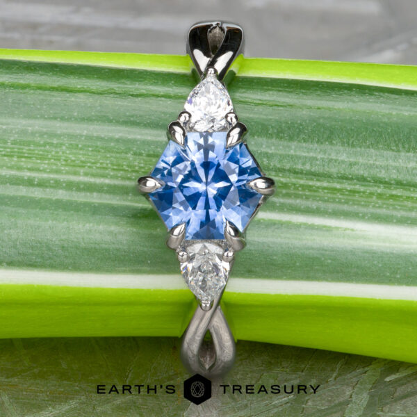 The "Elowen" ring in platinum with 1.09-Carat Montana Sapphire A pendant inspired by the "Elowen" ring in platinum, which features a 1.09-carat Montana sapphire at its center, flanked by two white pear-shaped stones, is elegantly set and displayed on a green striped leaf.