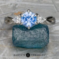 The "Elowen" ring in platinum with a 1.09-carat Montana Sapphire is displayed on a rough blue crystal, featuring a vivid blue square-cut center stone and two smaller clear stones. "EARTH'S TREASURY" appears at the bottom.