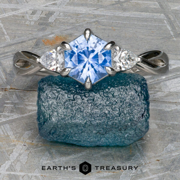 The "Elowen" ring in platinum with 1.09-Carat Montana Sapphire The "Elowen" ring in platinum with a 1.09-carat Montana Sapphire is displayed on a rough blue crystal, featuring a vivid blue square-cut center stone and two smaller clear stones. "EARTH'S TREASURY" appears at the bottom.