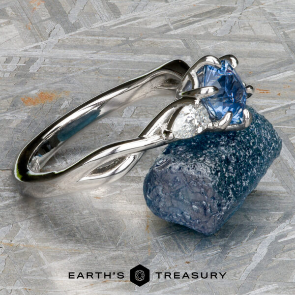 The "Elowen" ring in platinum with 1.09-Carat Montana Sapphire The "Elowen" ring in platinum with a 1.09-carat Montana sapphire rests on a rough blue mineral, showcasing the "Earth's Treasury" logo and text below.