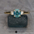 The “Stacked from 16 images. Method=B (R=1,S=3)” three-stone ring, featuring a teal oval gemstone and two clear side stones, is shown upright on a rough dark stone with the Earth’s Treasury logo below.