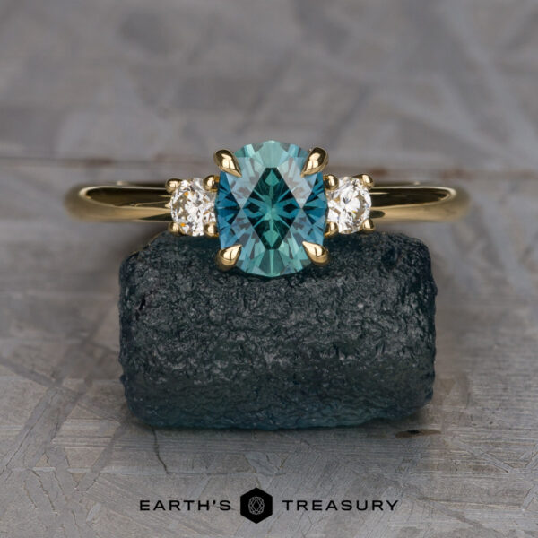The "Alocasia" ring in 18K yellow gold with 0.91-carat Montana sapphire The “Stacked from 16 images. Method=B (R=1,S=3)” three-stone ring, featuring a teal oval gemstone and two clear side stones, is shown upright on a rough dark stone with the Earth’s Treasury logo below.