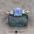 A platinum "Carina" ring with a 1.29-carat Ceylon sapphire and clear side stones is displayed on a textured blue-green block, with "EARTH'S TREASURY" written below.