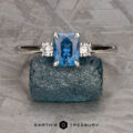 The "Alocasia" ring in 18K white gold features a 1.00-carat rectangular Montana sapphire and two round diamonds, shown atop a rough blue crystal with the "Earth's Treasury" logo below.