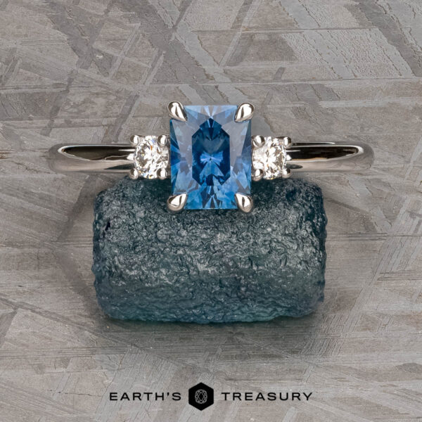 The "Alocasia" ring in 18K white gold with 1.00-carat Montana sapphire The "Alocasia" ring in 18K white gold features a 1.00-carat rectangular Montana sapphire and two round diamonds, shown atop a rough blue crystal with the "Earth's Treasury" logo below.