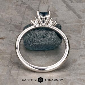 The "Alocasia" ring in 18K white gold features a 1.00-carat rectangular Montana sapphire and two round diamonds, shown atop a rough blue crystal with the "Earth's Treasury" logo below.