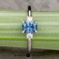 The "Alocasia" ring in 18K white gold features a 1.00-carat rectangular Montana sapphire and two round diamonds, shown atop a rough blue crystal with the "Earth's Treasury" logo below.