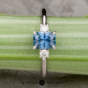 The "Alocasia" ring in 18K white gold features a 1.00-carat rectangular Montana sapphire and two round diamonds, shown atop a rough blue crystal with the "Earth's Treasury" logo below.
