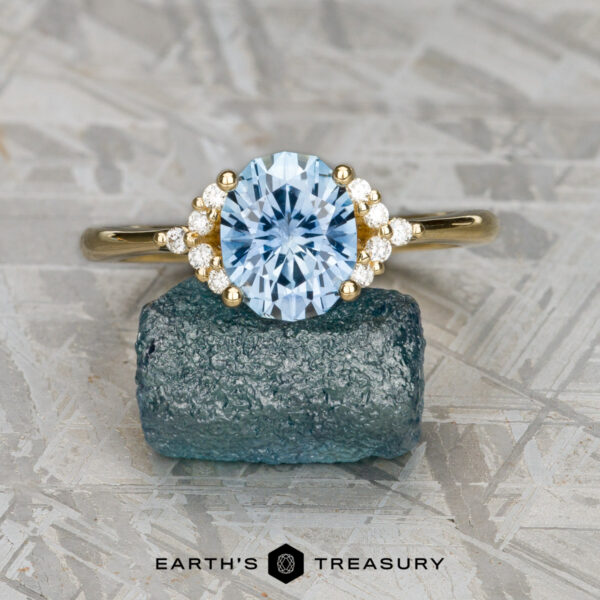 The "Chiron" ring in 18k yellow gold with 2.24-carat Montana sapphire The "Chiron" ring in 18k yellow gold, featuring a 2.24-carat Montana sapphire and diamond accents, rests on a rough blue stone above the text "EARTH'S TREASURY," capturing celestial elegance.