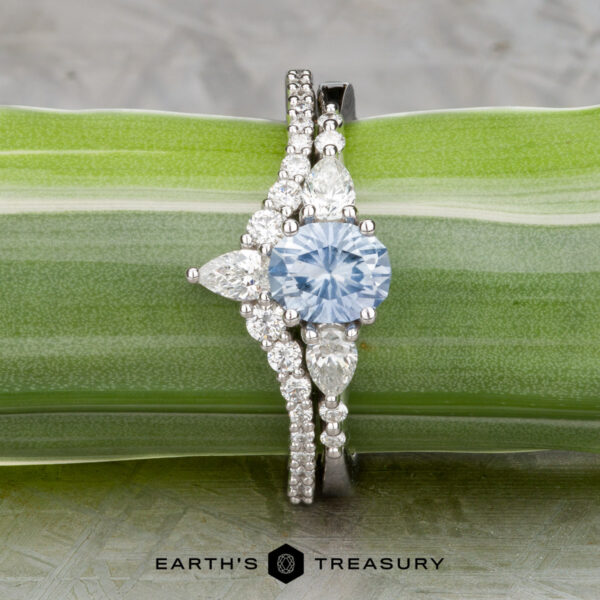 The "Carina" ring in 14k white gold with 0.95-carat Montana sapp The "Carina" ring in 14k white gold features a 0.95-carat Montana sapphire, accented by pear-shaped and round white stones, shown on a green leaf background.