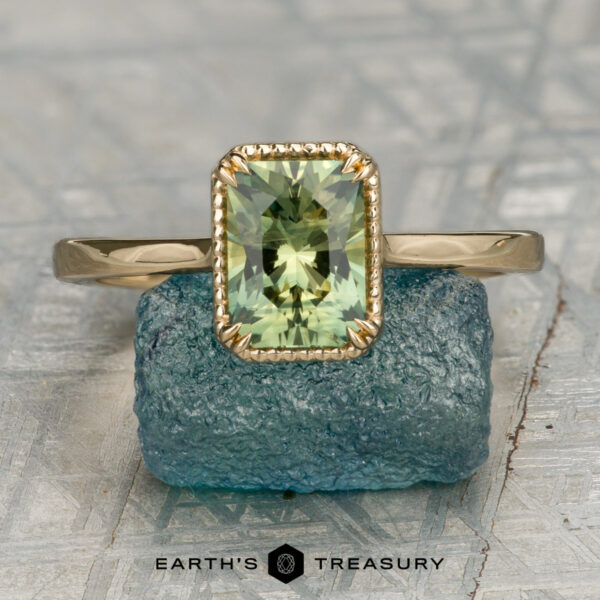 The "Ainsley" ring in 14K yellow gold with 1.54-carat Australian sapphire The "Ainsley" ring in 14K yellow gold, featuring a 1.54-carat Australian sapphire, is shown upright on a blue stone with the "Earth's Treasury" logo and text below.