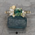 The "Ilana" ring in 14K yellow gold features a 1.06-carat Australian sapphire as its square centerpiece, accented by assorted side stones. It’s showcased on a rough blue stone with the EARTH'S TREASURY logo below, radiating elegance and beauty.