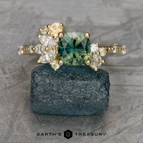 The "Ilana" ring in 14K yelow gold with 1.06-carat Australian sapphire The "Ilana" ring in 14K yellow gold features a 1.06-carat Australian sapphire as its square centerpiece, accented by assorted side stones. It’s showcased on a rough blue stone with the EARTH'S TREASURY logo below, radiating elegance and beauty.