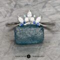 The "Selene" band in 18k white gold features marquise and round blue and clear gemstones in a double band jacket design, elegantly displayed standing on a rough blue crystal.