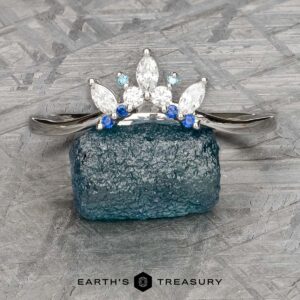 The "Selene" band in 18k white gold features marquise and round blue and clear gemstones in a double band jacket design, elegantly displayed standing on a rough blue crystal.