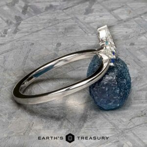The "Selene" band in 18k white gold, featuring a gemstone cluster setting and elegant Double Band Jacket design, is propped against a rough blue stone on a textured gray surface.