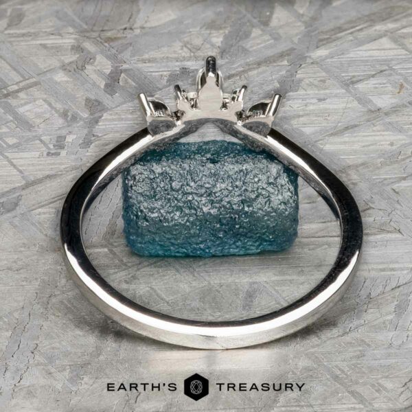 The "Selene" band in 18k white gold The "Selene" band in 18k white gold, featuring a rough blue gemstone and a Double Band Jacket, is photographed on a textured gray surface with the "Earth's Treasury" logo at the bottom.